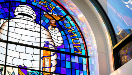 Stained glass window depicting the Bibendum figure holding a lamp with blue, purple, and yellow hues. Text appears around the upper and side edges. The image shows vivid colors and light streaming through the glass.