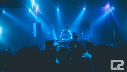 Two DJs stand behind a mixing console on a blue-lit stage, playing music for a crowd. The audience's silhouettes and raised hands are visible in front of the stage, showcasing the energetic atmosphere. The setting suggests a lively concert or nightclub event.