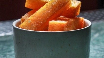 A white and gray ceramic bowl filled with thick, golden-brown fried yucca sticks. The bowl is set on a green textured surface with a dark background. The yucca sticks are crispy and appear freshly cooked, ready to be eaten.