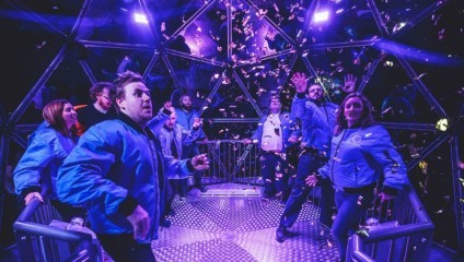 A group of people in blue jackets are inside a geodesic dome with a metal floor, participating in an activity where they are trying to catch floating confetti. The scene is brightly lit with a purple hue, creating an exciting and lively atmosphere.