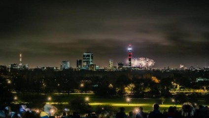 A nighttime cityscape featuring a brightly lit skyline with fireworks bursting near a tall structure. In the foreground, a crowd of people can be seen gathered on a grassy hill, enjoying the view. The scene is illuminated by streetlights scattered through the park.