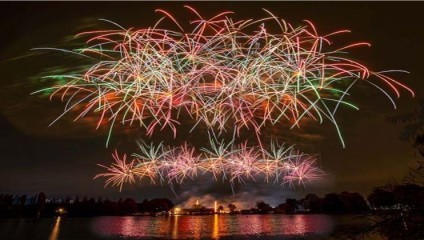 A nighttime scene with vibrant fireworks lighting up the sky above a calm body of water. Multiple bursts of red, green, yellow, and purple fireworks create a striking display, with reflections visible on the water’s surface. The landscape below is slightly illuminated.