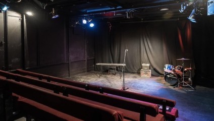 A dimly lit small theater stage with a microphone on a stand at the center, a keyboard on a stand to the left, and a drum set with two amplifiers to the right. The seating area consists of maroon-colored rows of chairs facing the stage.