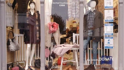 Storefront display featuring two mannequins in casual outfits, one in a dress with a scarf, the other in a shirt and jeans. Various clothing items and accessories are arranged inside, with donation signs and a Be Great. DONATE message visible.