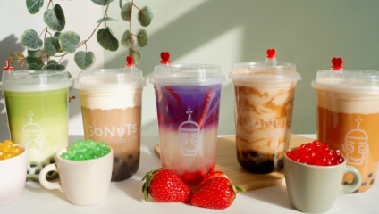 Five cups of colorful bubble tea, each with unique layers and toppings, are arranged in a row on a wooden surface. In front of the cups are small bowls of assorted boba pearls and fresh strawberries. A leafy plant is in the background, basking in sunlight.