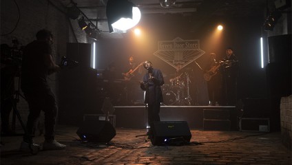 A band performs on a dimly lit stage at the Green Door Store, with a singer at the forefront holding a microphone. Stage lights are directed towards the singer, and a cameraman is filming the performance from the left side. Instruments and band members are in the background.