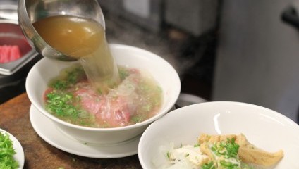 A ladle pours steaming hot broth into a bowl containing thinly sliced raw meat, fresh chopped green onions, and translucent noodles. Another bowl with tofu, green onions, and noodles is partially visible to the right. Fresh lettuce is seen in the corner.