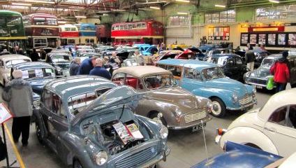 Inside the Transport museum. Rows of different coloured Morris Minors and old coloured buses in the background