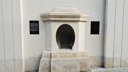 A small, arched stone enclosure embedded in a building's white exterior wall showcases a rock inside. Two black plaques with text are mounted on either side of the enclosure. The surrounding area has a light, polished stone base.