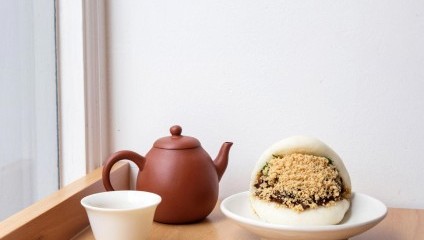 A white steamed bun filled with savory ingredients and topped with crushed peanuts sits on a wooden tray next to a small white cup and a brown teapot. The background is a white wall and window, providing a minimalistic and serene setting.