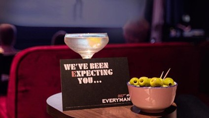 A cocktail glass with an olive garnish sits on a small table next to a bowl of olives. A black card with white text reads WE'VE BEEN EXPECTING YOU.... The scene is set in a dimly lit cinema with a blurred screen and audience in the background.