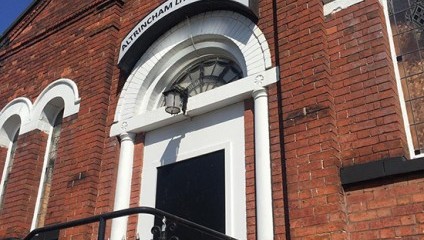 A brick building with arched windows and a door, marked by a white arch above it. A sign above the door reads Altrincham Little Theatre. Black metal railings lead up to the entrance. The sky is clear and blue.