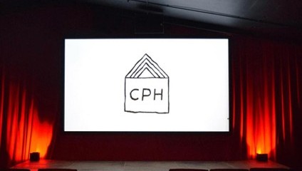 A dimly lit theater room with a glowing red ambiance. The theater screen displays a white background with a black line drawing. The drawing resembles an open book or layered shapes and the letters CPH are visible in the center.