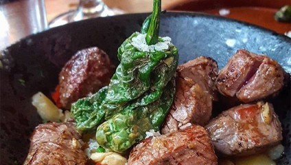 A close-up of a dish featuring juicy, grilled meat cubes topped with blistered green peppers and sprinkled with coarse salt. The food is served in a dark, rustic bowl, and the presentation highlights a hearty and savory meal.