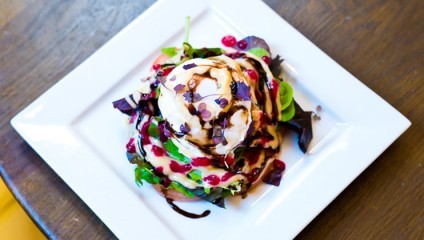 A gourmet salad is presented on a white square plate. The salad includes leafy greens, drizzled with creamy dressing, garnished with red pomegranate seeds, and accented with a balsamic glaze. A wood surface serves as the backdrop.