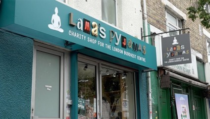 Front view of a shop named Lama's Pyjamas, a charity shop for the London Buddhist Centre. The storefront features colorful signage with a small Buddha figure, glass windows, and a door displaying an Open sign. Adjacent building walls are visible.