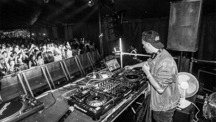 A DJ wearing a denim jacket and cap performs on stage at a concert. He is surrounded by DJ equipment and is using headphones to mix tracks. A large crowd in front of the stage dances energetically to the music. The atmosphere appears lively and enjoyable.
