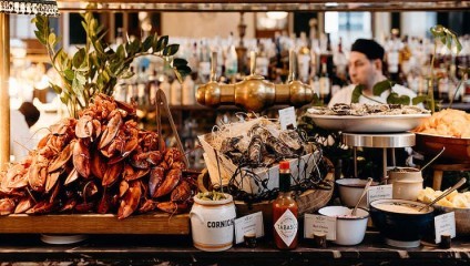 A seafood buffet is displayed featuring a large pile of cooked lobsters, oysters on a bed of ice, and various condiments including cornichons and Tabasco sauce. A bartender is visible in the background in front of a well-stocked bar.