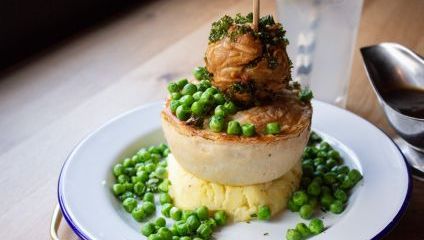 large pie on top of mash potato, gravy, and lots of green peas on top of and around the pie