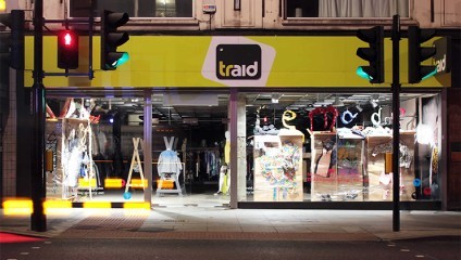 Storefront of a TRAID (Textile Reuse and International Development) shop at night, featuring a brightly lit interior showcasing clothing items. The store entrance is flanked by two mannequins and decorative elements. Traffic lights are visible outside.