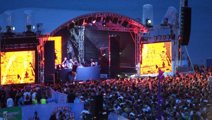 A large crowd enjoys a beachside concert at dusk, with performers on an elevated stage featuring vibrant, illuminated screens. The stage is equipped with an arched roof and various lighting rigs, while the audience revels under the evening sky.