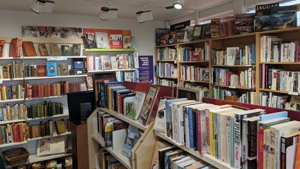 A cozy bookstore with shelves filled with books of various genres. The space is well-lit with track lighting, and there's a mix of contemporary and vintage books. Signage for different sections is visible, and a Wanted: Books and People sign is prominently displayed.