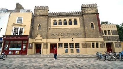 The image shows the exterior of the Pavilion Theatre, a historic brown brick building with a crenellated roofline. The entrance has several posters, and there is a ticket office on the left. Bikes are parked on the right, and a person is walking by.