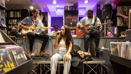 A band performs in a record store. A woman sits on stage, singing into a microphone. Behind her, two men play guitars while seated on amplifiers. The store's shelves are filled with vinyl records. Purple lighting illuminates the scene, and a banner reads album day.