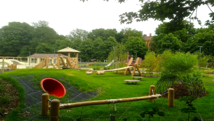 The Level Brighton A diverse playground with various equipment, including a red slide, wooden climbing structures, swings, and picnic tables. The area is surrounded by greenery and trees, giving it a natural, park-like feel. In the distance, a building is partially visible.