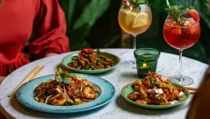 3 Bowls of Asian food on a white marble table with 2 tall cocktail glasses - one red and one yellow each with ice and straws