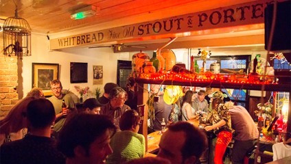 A cozy, crowded pub with wooden decor lit by warm, dim lights. People are mingling and chatting around the bar area, which is decorated with bright string lights and various quirky ornaments. A sign above reads WHITBREAD Fine Old STOUT & PORTER.