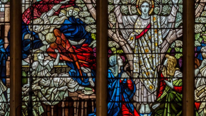 A detailed stained glass window depicts a biblical scene with three women in vibrant robes and a radiant angel with a golden halo. Intricate patterns and an array of bright colors showcase symbolism and artistry, with trees and other figures filling the background.
