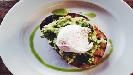 A plate with a slice of toasted bread topped with mashed avocado and a poached egg. The dish is garnished with a drizzle of green sauce around the perimeter of the plate.