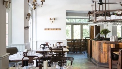 A cozy restaurant interior featuring wooden tables set with glassware and white napkins. The space has a rustic charm with wooden floors, a wooden bar, and soft lighting from wall-mounted lamps and chandeliers. Large windows let in natural light.