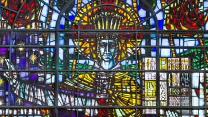 A vibrant stained glass window depicting a central figure with a halo, arms outstretched. Surrounding the figure are colorful geometric patterns and text fragments that read, I am the life, Behold I am alive forever more, amidst other intricate designs.
