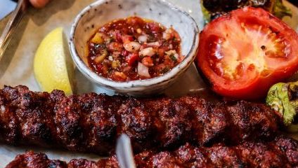 two meat kebabs on skewers lined up, a tomato, lemon slice and red dipping sauce