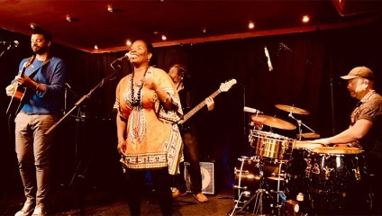 A band performs on stage in a dimly lit venue. The vocalist, wearing a colorful patterned dress, sings into the microphone, while the guitarist, bassist, and drummer play their instruments. The black background features subtle lighting and a few spotlights.