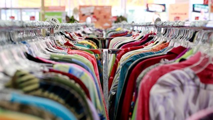 A close-up of multiple racks filled with colorful clothing items on hangers in a store. The perspective is from the middle, looking down the rows of clothes. The background shows a brightly lit space with partial visibility of signs and shelves.