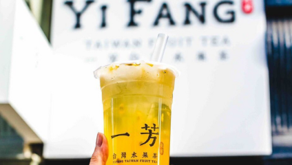 A hand holding a clear cup of green tea topped with foam, with a white straw sticking out. The cup has yellow and black Chinese characters and the text “YI FANG TAIWAN FRUIT TEA.” The sign for Yi Fang is blurry in the background.