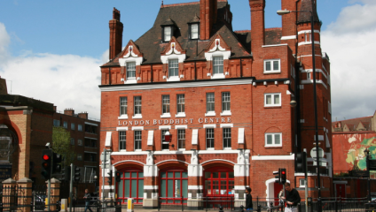 A large red-brick building with white accents, displaying the words London Buddhist Centre on the front. It has a historic architecture with tall windows and several chimneys. The surrounding area includes traffic lights, pedestrians, and nearby buildings.
