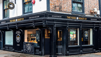 Street view of The York Roast Co. corner shop, a black-painted establishment with large windows revealing the interior. Signage on the exterior lists Eat In or Take Out and Serving the World Famous Yorky Pud among other details.