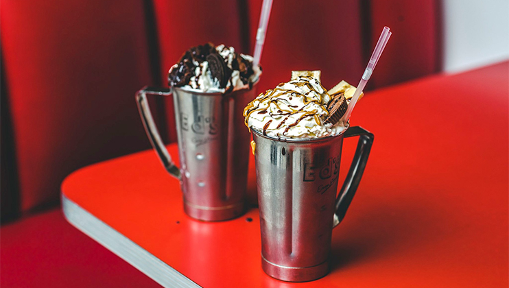 Two metal milkshake cups on a red table, each topped with whipped cream and drizzled with chocolate sauce. One milkshake is garnished with chocolate chunks, and the other with caramel and small wafer pieces. Red cushioned seating is visible in the background.