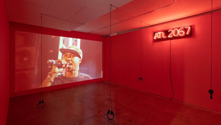 A red-lit room features a large screen displaying a person holding a microphone and wearing a cap. Four sets of black headphones hang from the ceiling on thin cords. A neon sign on the wall reads ATL 2067.