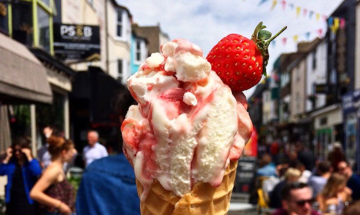 A close-up of a strawberry-topped ice cream cone with pink and white swirled ice cream. The background features a busy, vibrant street scene with buildings, colorful bunting, and people enjoying outdoor activities.