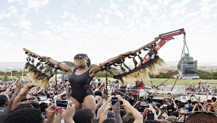 A performer wearing a black outfit and a mask holds a large feathered prop resembling wings, standing amidst a crowd. In the background, a crane is removing a statue. The crowd is watching the scene, with some people taking photos and videos.