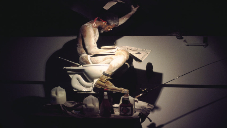 A man covered in white paint, wearing only boots and a red headband, sits on a toilet reading a newspaper. The scene is dramatic with dark lighting. Nearby items include milk, ketchup, mustard, and a fishing rod extending from the toilet.