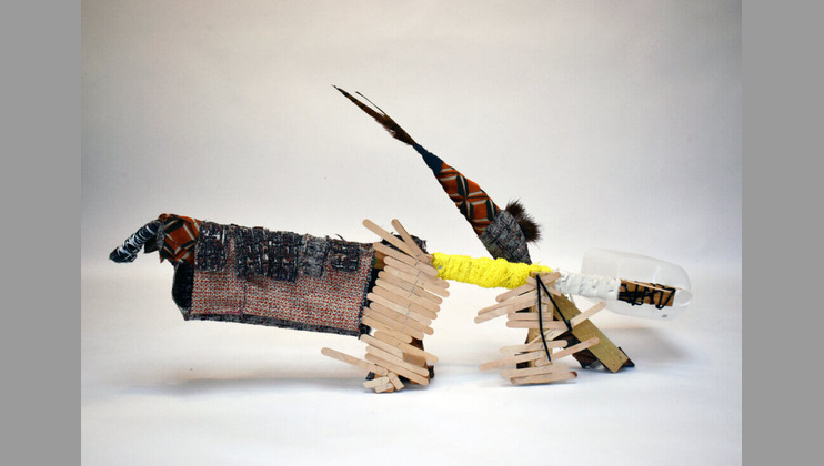 A whimsical sculpture resembling a dog made from assorted materials, including popsicle sticks, fabric, feathers, and a yellow knitted item. The elongated body and angular legs contribute to its playful, abstract design, set against a neutral background.