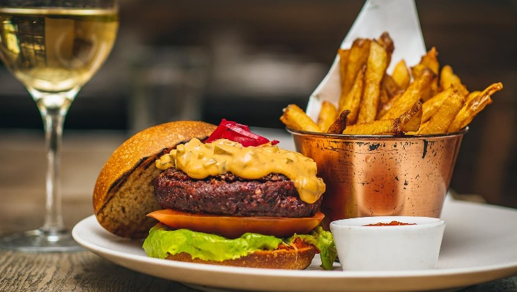 A gourmet cheeseburger with lettuce, tomato, and pickles is served on a white plate alongside a metal cup of golden french fries. A small bowl of sauce sits on the plate, and a glass of white wine is visible in the background.