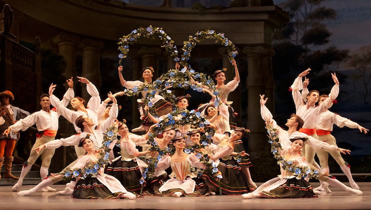 A group of ballet dancers pose on stage, holding floral wreaths in the air and wearing traditional costumes with puffy white sleeves and dark skirts or pants. The backdrop features classical architecture and painted scenery, creating a theatrical atmosphere.