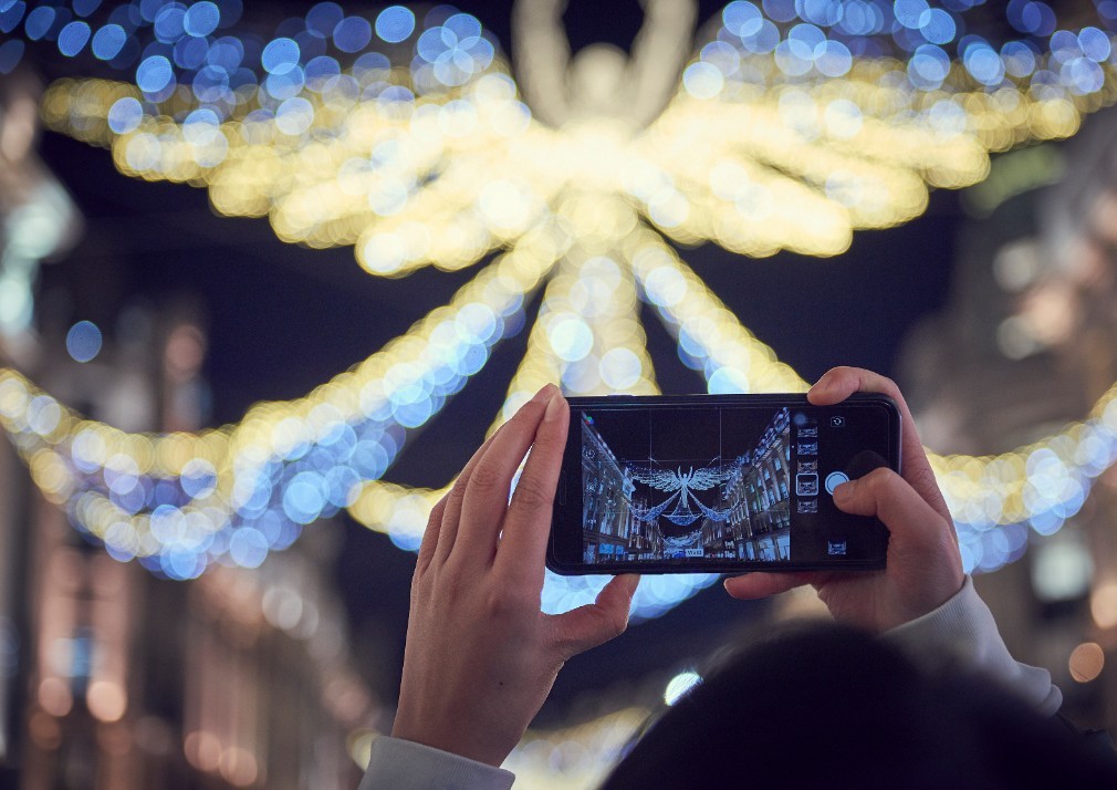 A person holds a smartphone to capture a photo of vibrant, festive lights shaped like an angel hanging above a street at night. The background is adorned with blue and yellow bokeh lights creating a magical atmosphere.